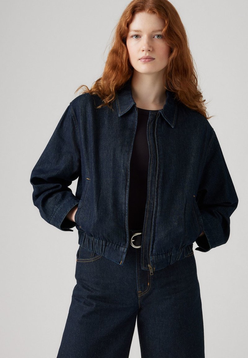 Veste en denim noir avec taille élastique, col évasé et fermeture éclair à l'avant. Dotée de poches latérales et d'un design à coupe décontractée.