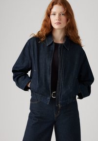 Tamno traper jakna opuštenog kroja, s ovratnikom, elastičnim strukom i dva prednja džepa. Ima vidljive šavove i patentni zatvarač.