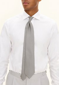 Gray patterned tie with a diamond design, made of smooth fabric, worn over a white shirt with a classic collar and light gray trousers.