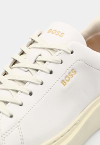 Witte lederen sneaker met crèmekleurige veters en een beige zool. Voorzien van het "BOSS"-logo in goud aan de zijkant. Glad van textuur en een minimalistisch ontwerp.