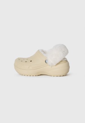 Sabot beige avec une douce doublure en fourrure blanche et des trous de ventilation, présentant une forme arrondie et des accents texturés sur le dessus et la semelle.