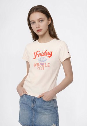 Млада жена, носеща кремава тениска с текст "Friday Noodle Club" и графика на купа с юфка, съчетана със синя дънкова пола.