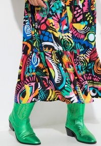 Grüne metallische Cowboystiefel mit spitzen Zehen und dekorativer Stickerei, kombiniert mit einem mehrfarbigen, abstrakt gemusterten Kleid.