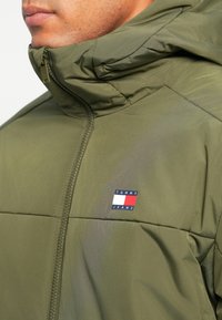 Green puffy jacket with a high collar, featuring a visible zipper and a logo patch in red, white, and blue on the left chest.