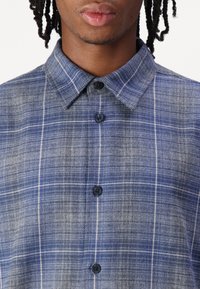 Camicia di cotone a quadri blu e grigia con colletto classico, caratterizzata da bottoni scuri e un tessuto testurizzato con motivo a griglia bianca.