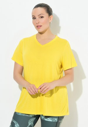 T-shirt giallo a V in tessuto morbido, con maniche corte e vestibilità rilassata, abbinato a leggings a fantasia in tonalità verdi e grigie.