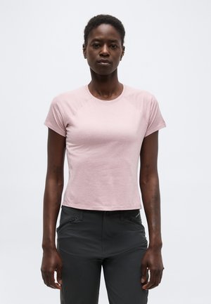Kvinde med kort hår iført en lys pink, tætsiddende t-shirt med korte ærmer og mørkegrå bukser, stående mod en ensfarvet baggrund.