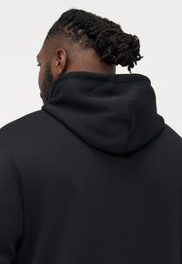 Schwarze Kapuzenjacke mit weicher Textur, großer Kapuze und einem schlichten, minimalistischen Design. Zeigt eine verkürzte Sicht von hinten.