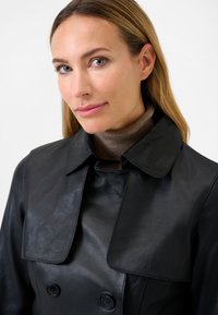 Black leather trench coat with a wide collar, double-breasted front, and smooth texture. Underneath, a brown sweater is partially visible.