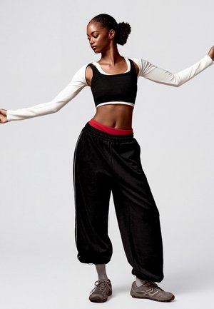 Jeune femme aux cheveux attachés en chignon pose avec les bras étendus, portant un crop top noir, des manches longues blanches, un pantalon noir ample et des baskets grises.