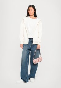 Crème zip-up hoodie, witte graphic t-shirt, wijdvallende blauwe denim jeans, witte sneakers en een kleine roze handtas met hardware accenten.