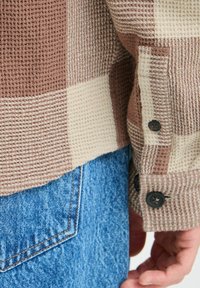 Checkered shirt featuring brown and cream squares, textured fabric, and buttoned cuffs. Paired with blue denim jeans with a back pocket.