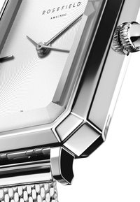 Montre en acier inoxydable avec un boîtier octogonal, un cadran texturé blanc, des aiguilles argentées et une couronne détaillée. Le bracelet en maille ajoute une touche raffinée.