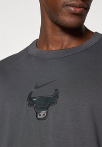 Grå Nike T-skjorte med en mørk, brodert Chicago Bulls-logo på brystet, med rund hals og teksturert stoff.