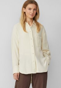 Crèmekleurig button-up shirt van textuurstof, met een voorzak, lange mouwen en een gebogen zoom. Gecombineerd met bruine broek.