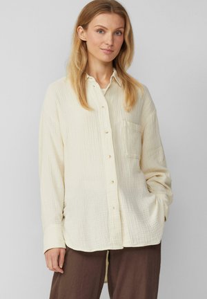 Crèmekleurig button-up shirt van textuurstof, met een voorzak, lange mouwen en een gebogen zoom. Gecombineerd met bruine broek.