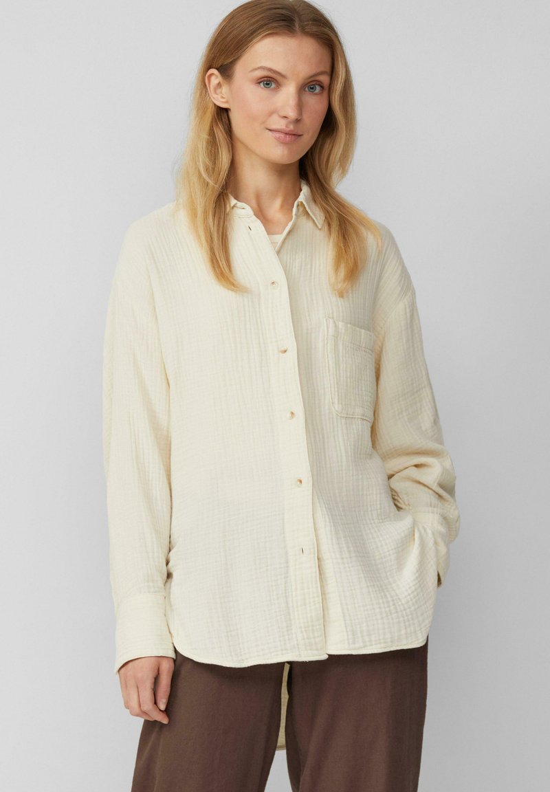 Crèmekleurig button-up shirt van textuurstof, met een voorzak, lange mouwen en een gebogen zoom. Gecombineerd met bruine broek.