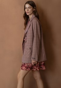 Blazer de cuadros marrón con grandes solapas y dos bolsillos frontales, llevado sobre una falda floral rosa con capas. Textura suave y ajuste entallado.
