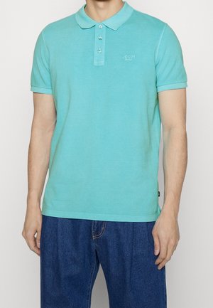 Mand iført en lyse turkis kortærmet poloshirt med tre knapper og mørkeblå denimjeans, stående med armene afslappet.