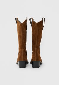 Ζευγάρι καφέ σουέντ μπότες μέχρι το γόνατο με μαύρα χοντρά τακούνια, εμφανιζόμενες από πίσω σε ουδέτερο γκρι φόντο.