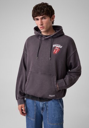 Giovane uomo con capelli ricci indossa una felpa con cappuccio Rolling Stones grigio scuro e jeans blu, in piedi davanti a uno sfondo grigio chiaro uniforme.