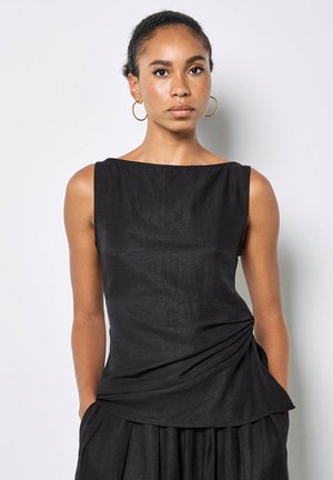 Femme aux cheveux tressés et boucles d'oreilles créoles en or, portant un haut noir sans manches avec des fronces sur les côtés, debout devant un fond blanc uni.