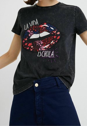 Schwarzes ausgebleichtes T-Shirt mit großem rotem Blumenlippen-Design und weißem Text „La Vida Es Chula“, kombiniert mit dunkelblauer Hose.