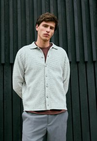 Helle graue, strukturierte Strickjacke mit durchgehender Knopfleiste und Kragen. Über einem dunklen Hemd getragen, kombiniert mit hellgrauen Hosen. Strukturierter Hintergrund.