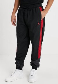 Zwarte windbreakerbroek met rode zijstrepen, elastische tailleband en boorden, met een gestructureerd zwart-grijs gedeelte bij de enkels.