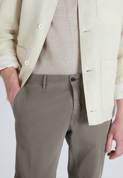 Giacca beige chiaro, abbottonata e a un petto, sopra una camicia a righe grigio chiaro. Pantaloni verde oliva con chiusura a bottone e tasche.
