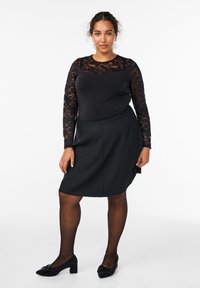 Ženska stoji v črni čipkasti dolgi srajci z dolgimi rokavi, črni krilu, prozornih črnih nogavicah in črnih čevljih na blok peto z mašnjami.