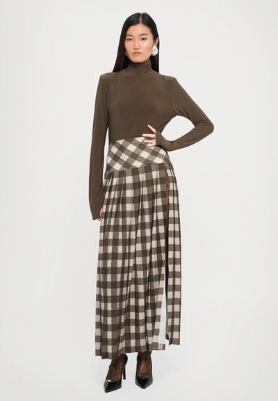 Brūna garām piedurknēm turtlenecks blūze, kas valkāta kopā ar kvadrātu svārkiem maksi garumā brūnos un bēšos toņos, kurus rotā ieloces un sānu šķirba. Melni augstpapēžu zābaki.