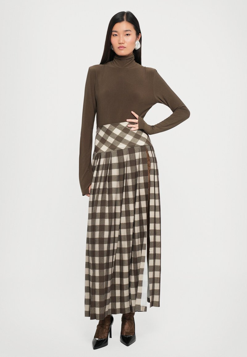 Top de punto marrón de mangas largas y cuello alto combinado con una falda larga a cuadros en tonos marrones y beiges, con pliegues y una abertura lateral. Botas de tacón negras.