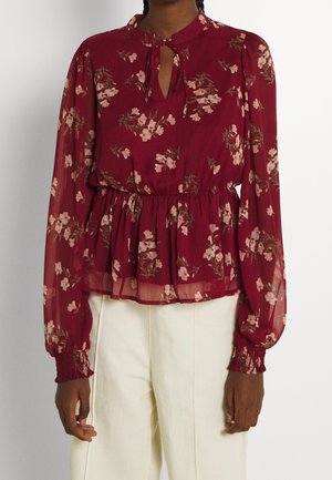 Donna che indossa una blusa floreale bordeaux a maniche lunghe con scollatura a chiave e pantaloni crema a vita alta.