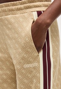 Χέρι με σκούρο δέρμα στην τσέπη μπεζ παντελονιών Guess με μοτίβο μονόγραμμα και μπορντό και κρεμ ρίγες στο πλάι.