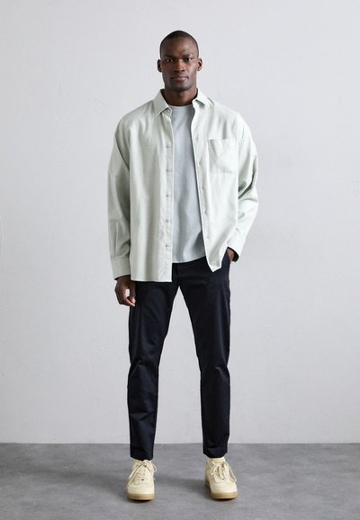 Camisa de botones verde claro sobre una camiseta gris claro, pantalones oscuros y zapatos beige. La camisa tiene un bolsillo, un corte relajado y una textura suave.