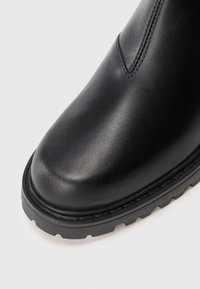 Close-up da ponta de uma bota de couro preta com detalhes em costura e sola de borracha textura grossa, em um fundo branco.