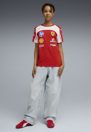 Giovane in piedi che indossa una maglia da corsa Ferrari rossa, jeans chiari e larghi e sneakers Puma rosse, su uno sfondo grigio uniforme.