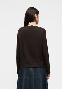 Γυναίκα με μακριά σκούρα μαλλιά που φοράει ασημένια κρίκους σκουλαρίκια, μακρυμάνικη καφέ μπλούζα και σκουρόχρωμη μπλε τζιν φούστα, γυρισμένη πλάτη σε λευκό φόντο.