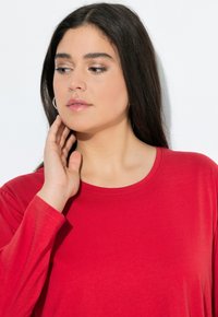 Červená košile s dlouhými rukávy a kulatým výstřihem, vyrobená z měkké tkaniny. Modelka si lehce dotýká obličeje, čímž předvádí volný střih košile.