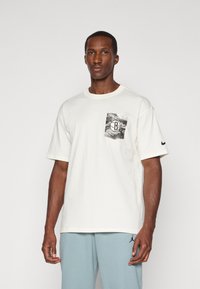 T-shirt en coton blanc avec un design de poche graphique noir et gris, col rond, manches courtes et un logo Nike noir sur la manche gauche.