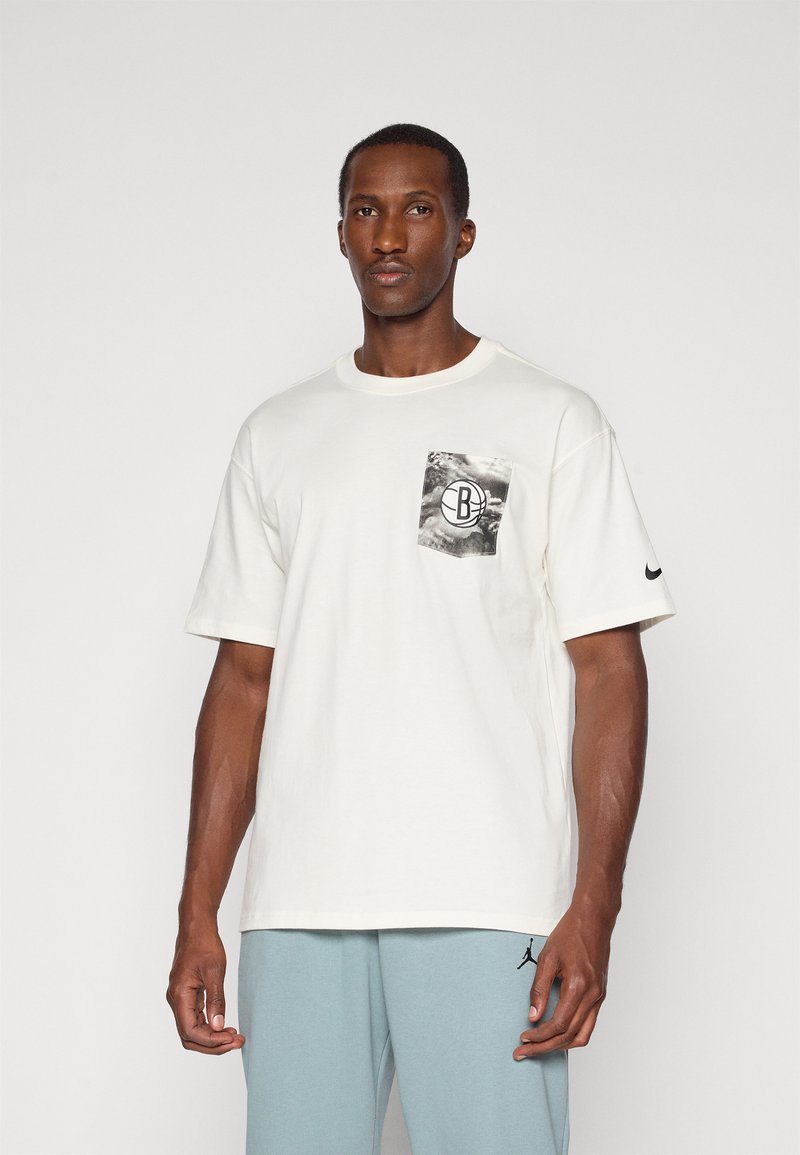 T-shirt en coton blanc avec un design de poche graphique noir et gris, col rond, manches courtes et un logo Nike noir sur la manche gauche.