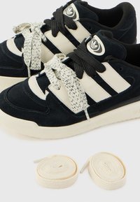 Black and beige sneaker made of suede and leather, featuring textured laces, three stripes, and two additional cream laces beside.
