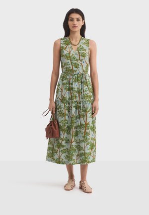 Femme portant une robe verte sans manches avec un imprimé d'arbres, tenant un sac à main marron et portant des chaussures tissées beiges, debout devant un fond uni.