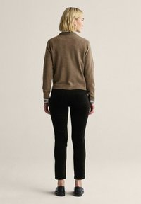 Brun tröja med vita och svarta randiga manschetter, i kombination med svarta byxor; modellen står med ryggen mot betraktaren, kort hår synligt.