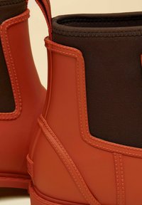 Stivaletti impermeabili con esterno in gomma rossa, fodera in tessuto marrone e dettagli cuciti lungo i bordi e le cuciture.