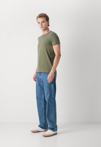 Olivgrünes kurzärmliges T-Shirt mit Rundhalsausschnitt, kombiniert mit hellblauen Jeans. Weiße Sneakers mit beige Akzenten runden das Outfit ab.