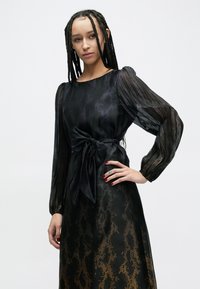 Schwarzes Kleid mit langen, transparenten Ärmeln, rundem Halsausschnitt und einer gebundenen Taillendetail. Es verfügt über ein strukturiertes Muster mit goldenen Akzenten, die in Schwarz übergehen.