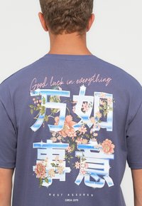 Persona con cabello corto y rizado lleva una camiseta azul con diseños florales y un texto que dice "Buena suerte en todo" y "Descansa tranquilo".