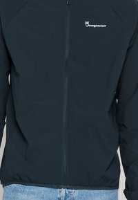 Tmavě modrá zipová bunda vyrobená z měkké látky, s kulatým límcem a decentním logem na levé straně. Lehká a hladká textura.